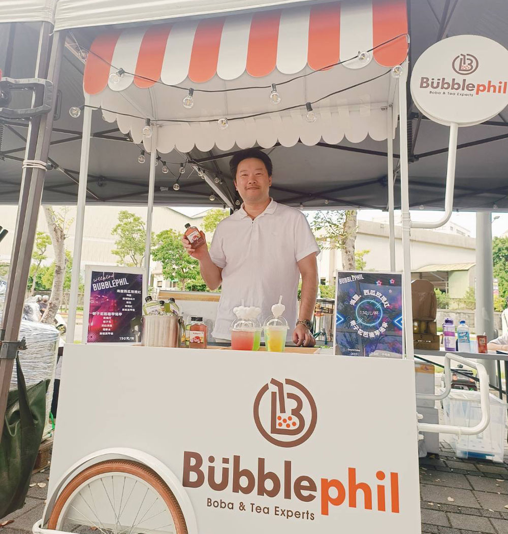 A smiling male brand representative standing inside a professional, white-and-orange striped BubblePhil mobile beverage kiosk showcasing product samples at an event.